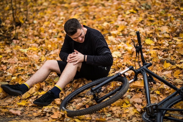 Free photo active young man holding by his hurt or broken leg while lying on autumn forest path by his bicycle