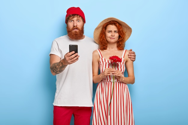 Free photo addicted puzzled bearded ginger man holds mobile phone and embraces girlfriend