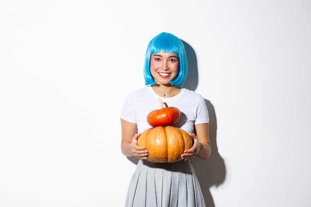 2つのかわいいカボチャを保持し、カメラに微笑んで、ハロウィーンパーティーのための女子高生の衣装を着て、青いかつらの愛らしいアジアの女性。