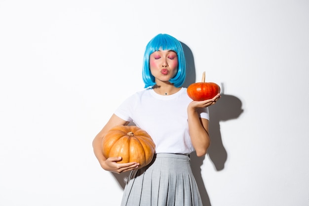 Adorable asian woman holding two pumpkins while kissing someone with closed eyes, celebrating halloween in blue wig and schoolgirl costume.
