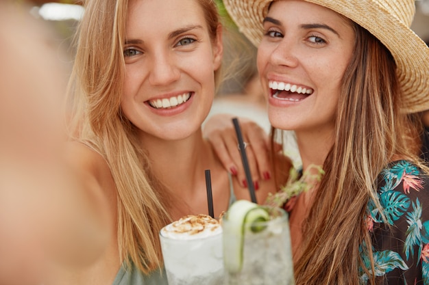 Adorable beautiful lesbian females have positive expressions, make selfie with unrecognizable electronic device, stand close to each other, demonstrate good relationships and love. Rest concept