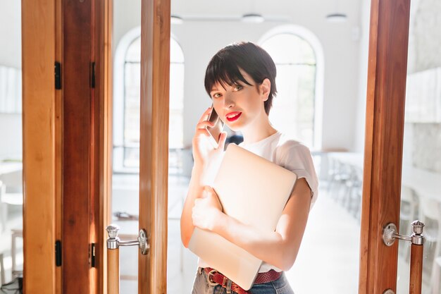 Adorable black-haired girl holding modern computer speaking on phone and cute smiling near office