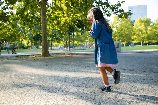 Adorable black haired little girl playing hopscotch in city park. Full length, copy space. Childhood concept