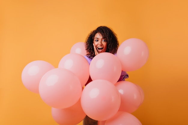 Прелестная черная женщина наслаждаясь партией с улыбкой. Очаровательная женская модель с розовыми гелиевыми шарами, стоящими на апельсине.