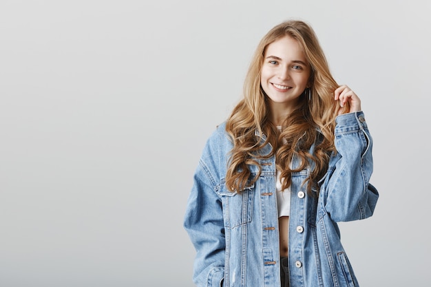 Очаровательная белокурая девушка улыбается счастлива в джинсовой куртке, стоящей над серой стеной
