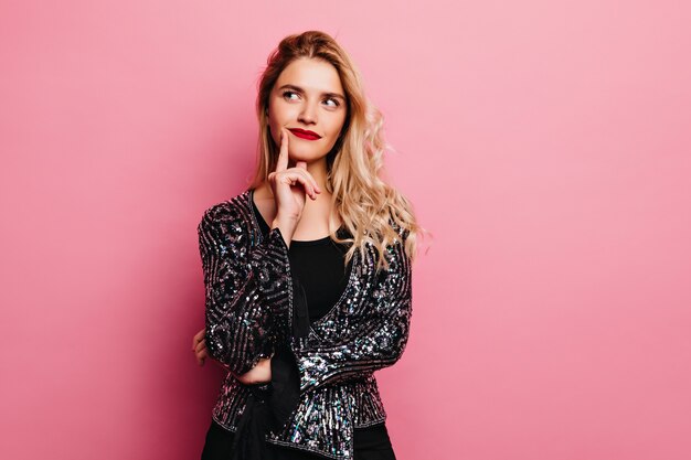 Adorable blonde girl posing with pensive face expression. Pretty young woman in sparkle jacket standing on pink wall.