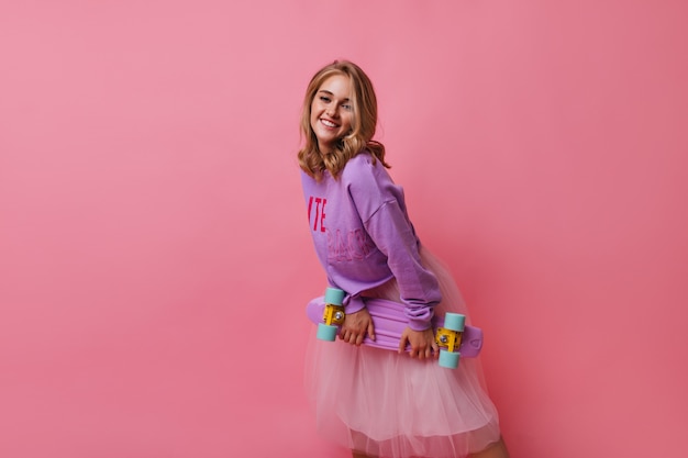 Adorable blonde girl in purple shirt posing. Blithesome curly female model in white skirt holding skateboard.