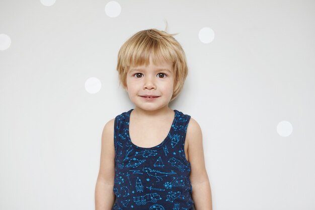 adorable blonde male toddler with fair hair and beautiful brown eyes with smile, standing against white wall with copy space for your promotional content