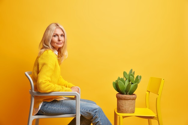 adorable blonde middle aged lady in casual clothes sits on chair opposite pot of cactus