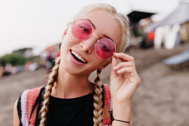 Adorable blonde woman posing at beach and touching her pink glasses.