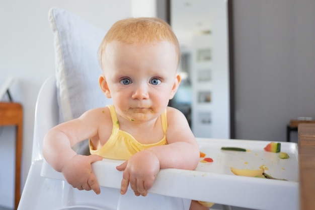 ハイチェアに座ってカメラ目線の顔に緑のピューレと愛らしい青い目をした女の赤ちゃんが汚れます。クローズアップショット。摂食プロセスまたは育児の概念