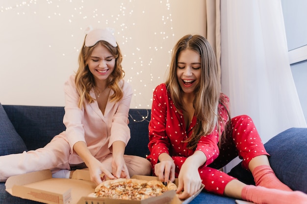 Adorable brunette girl in cute socks eating pizza in morning. Indoor photo of two ladies posing during breakfast in pajamas.