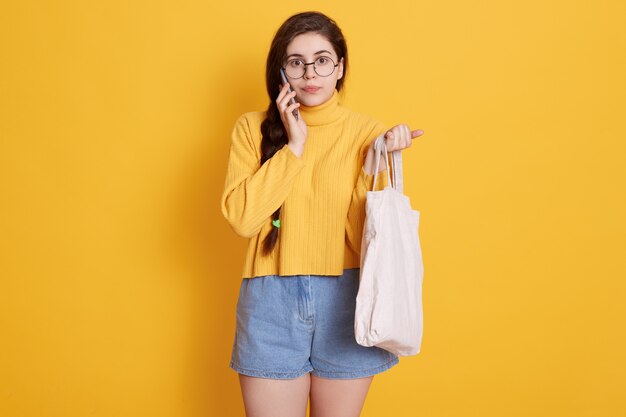 Adorable brunette woman wearing yellow sweater and short, holding bag in hand, talking to her friend via modern smart phoneç