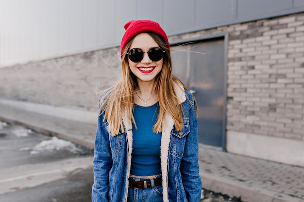 Очаровательная кавказская девушка в черных очках стоит на улице и улыбается. Привлекательная белая женщина в джинсовой куртке гуляет в весенний день.