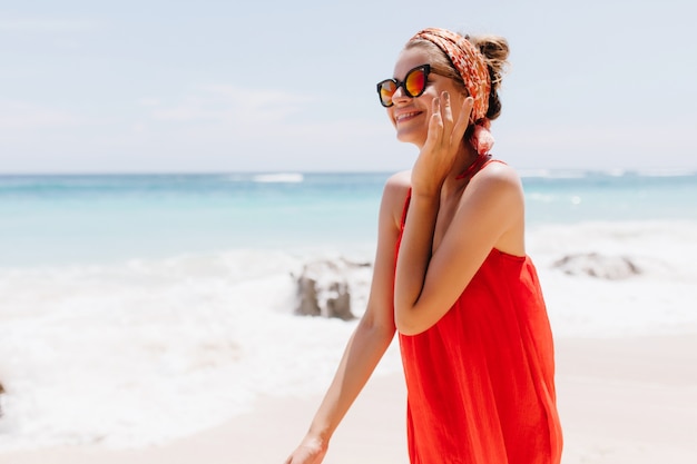 海の近くのエキゾチックな場所で夏を過ごす愛らしい白人の女の子。ビーチでポーズをとってサングラスで優雅な笑顔の女性の屋外写真