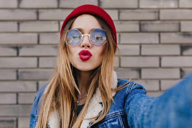 Adorable caucasian girl with straight hairstyle posing with kissing face expression on brick wall. Outdoor shot of blissful white lady in glasses and red hat expressing love while making selfie.