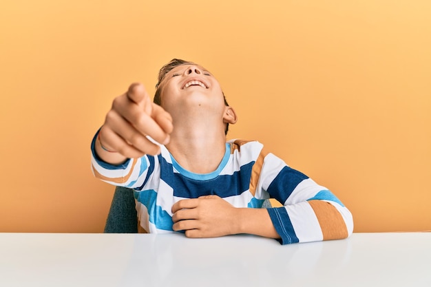 Free photo adorable caucasian kid wearing casual clothes sitting on the table laughing at you, pointing finger to the camera with hand over body, shame expression