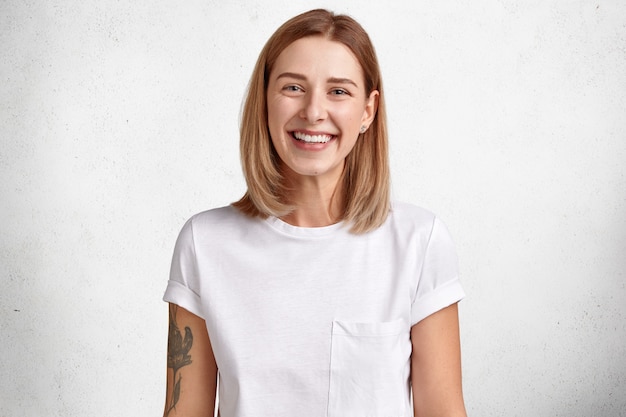 adorable caucasian woman with positive expression, has white perfect teeth, healthy skin, tattoo on arm, short hair rejoices good news