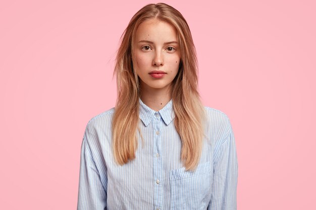 Adorable Caucasian young female with confident serious look, has long hair, dressed in stylish shirt, stands against pink wall, ready to talk with coworker.