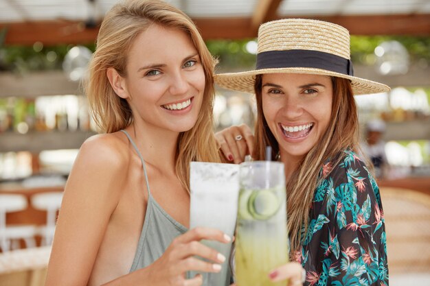 愛らしい陽気な女性は夏の帽子、ブラウスを着て、広い笑顔を輝かせ、カクテルのグラスをチャリンと鳴らし、夏休みの始まりを祝い、熱帯の国で休む機会を持つことを喜ぶ