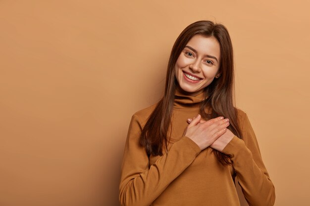 Adorable cheerful woman keeps both palms pressed together near heart in gratitude symbol