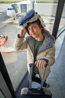 Очаровательный ребенок-пассажир в очках и чемодане, стоящий в аэропорту