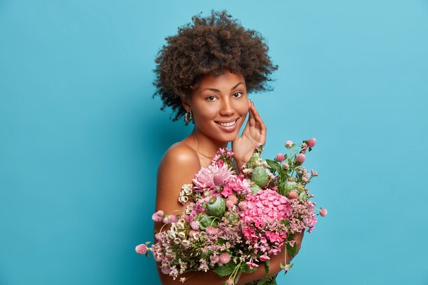 Adorable curly haired woman stands with bare shoulders touches face gently holds big beautiful bouquet of flowers expresses positive emotions