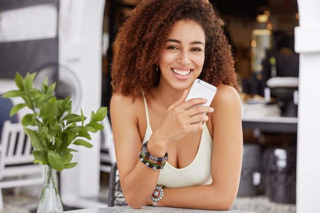 Adorable curly woman with positive expression holds cell phone in hands, messages in social networks, enjoys high speed internet connection in cafeteria.