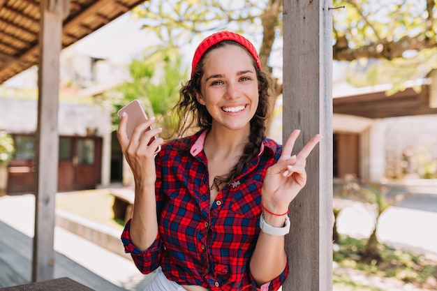 Adorable cute young woman with happy charming smile with smartphone rests outside in sunlight and shows peace sign. Hipster lifestyle, summer day