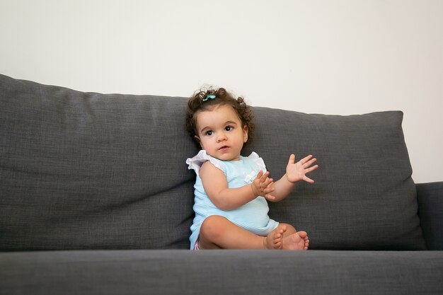 Очаровательная темная кудрявая девочка в бледно-голубой ткани сидит на сером диване у себя дома, смотрит в сторону и хлопает в ладоши. Ребенок дома и концепция детства