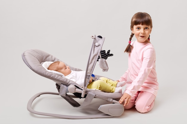 Бесплатное фото Очаровательная темноволосая девочка с косичками играет с новорожденной сестрой или братом, лежащим в кресле-вышибале
