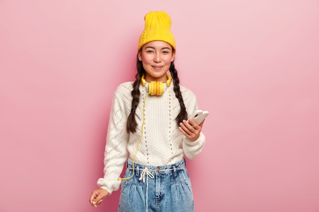 Adorable dark haired girl with pigtails, uses mobile phone for surfing social networks, wears stylish headgear