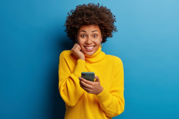 Adorable dark skinned adult woman dressed in yellow jumper using mobile phone with a happy expression