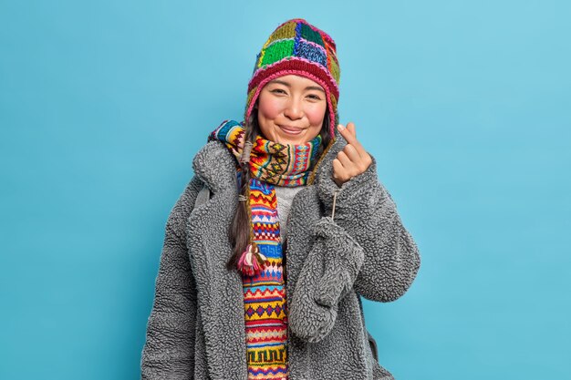 Adorable eastern Asian girl dressed in winter clothing makes korean like sign shows mini heart expresses love going to have walk poses against blue studio wall