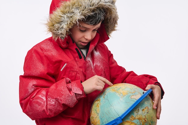 旅行先​を​探して​地球​に​指​を​向けて​、​フード付き​の​暖かい​雪​に​覆われた​赤い​ダウン​ジャケット​を​着た​愛らしい​小学生​の​白人​の​男の子​。​コピー​広告​スペース​と​冬​の​観光​コンセプト