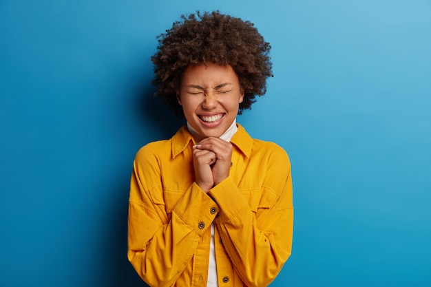 Adorable ethnic woman smiles toothily, keeps hands under chin, closes eyes, expresses happiness, dressed in stylish outfit, has polite calm expression, isolated over blue background