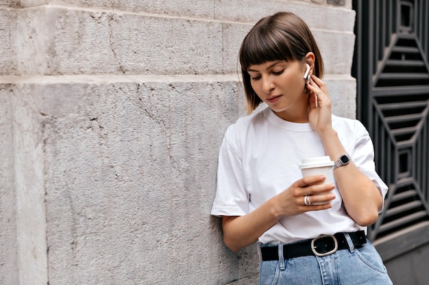 Очаровательная европейская девушка с короткими волосами в футболке и джинсах слушает музыку в беспроводных наушниках и пьет кофе на улице возле бежевой стены в городе