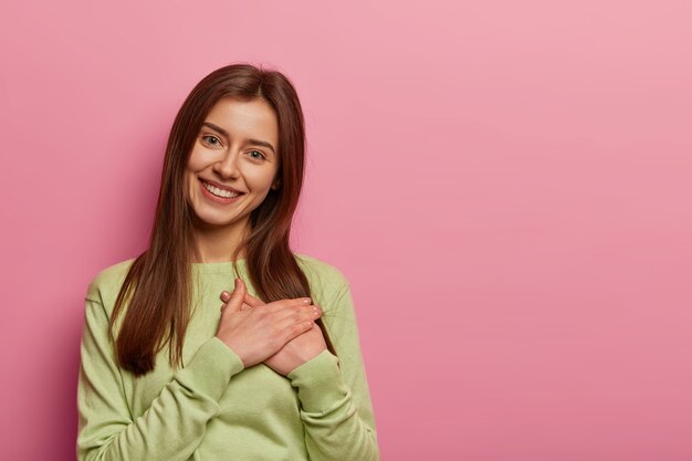 Adorable European woman pressed hands to palm, sees heartwarming scene, looks with admiration and gratitude, smiles gently