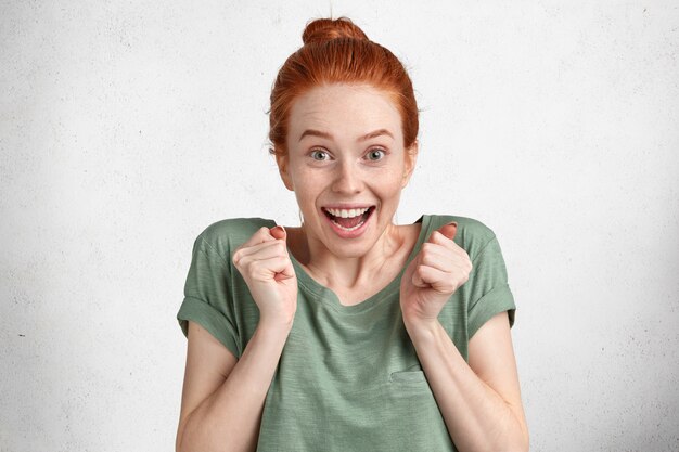 Adorable excited female with overjoyed expression, rejoices her success, keeps fists raised, dressed in casual clothing, isolated over white studio