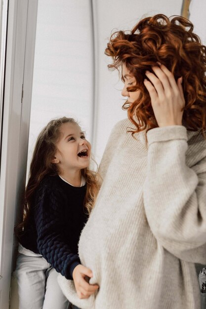 Adorable excited little girl is sitting near the window and looking up on her mother Pregnant woman in knitted pullover is playing with her little daughter in sunny day