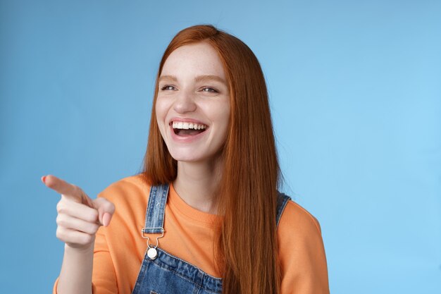 사랑스럽고 매력적인 젊은 빨간 머리 소녀는 즐겁게 웃고 있는 재미있는 광고를 보고 왼쪽을 가리키며 즐겁게 웃고 있습니다. 파란색 배경.