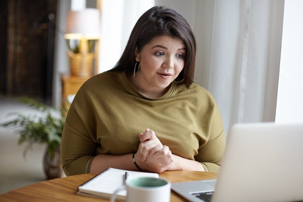 Adorable fashionable young plus size woman sitting at cozy cafeteria in front of open laptop, using free wifi while chatting online with her friend via video call, having excited look. Film effect