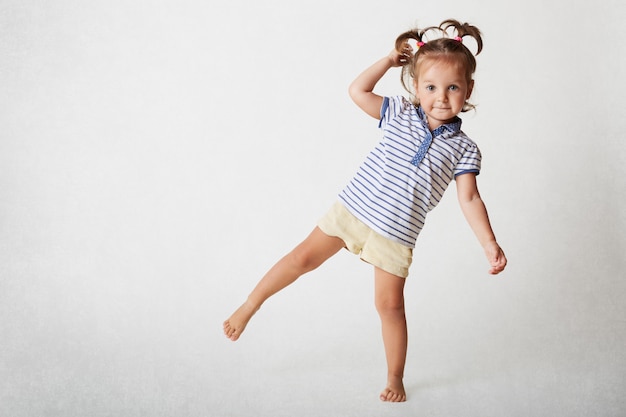 Adorable female child has funny expression, two pony tails, wears casaul t shirt and shorts, stands on one leg