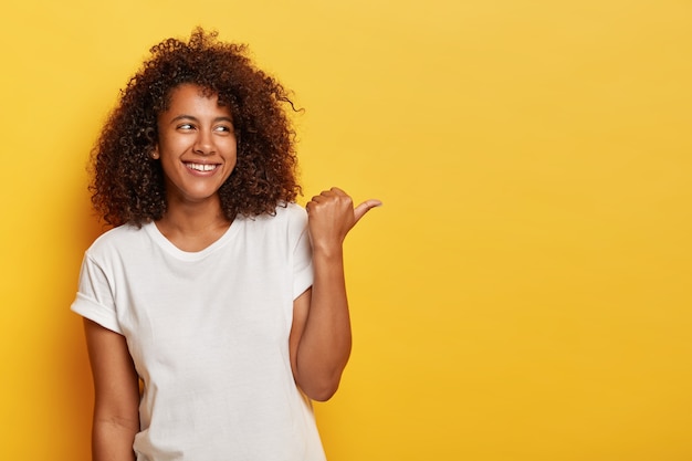 Adorable female student with bushy curly hair points thumb right, feels happy and relaxed, wears white casual t shirt, has sincere smile on face, isolated on yellow wall, shows something interesting