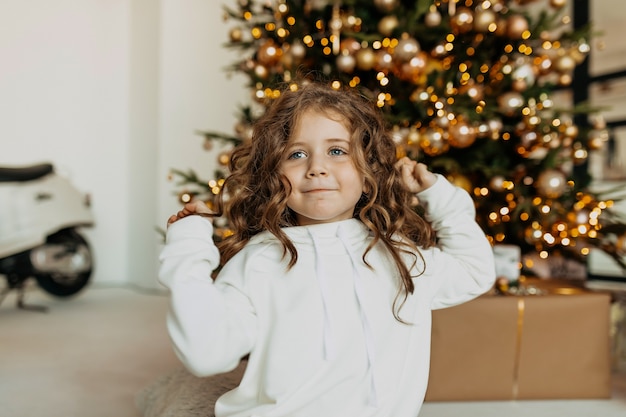 Adorable funny little girl dressed white clothes having fun in front of Christmas tree