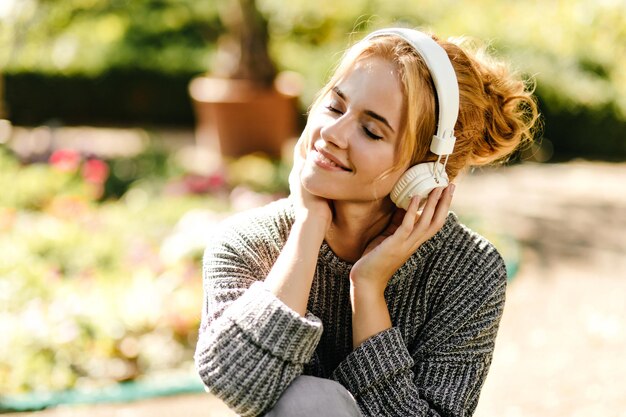 Adorable girl in gray knitted sweater listens to music with headphones on and sits in park