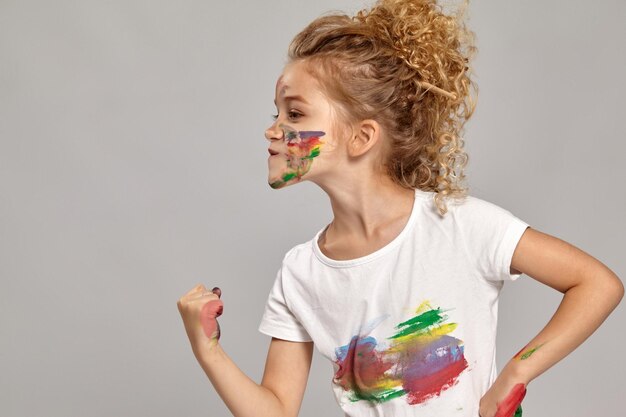 Adorable girl having a brush in her lovely haircut, wearing in a white smeared t-shirt. She is posing with a painted hands and cheeks, showing a fist to someone and looking angry, on a gray background