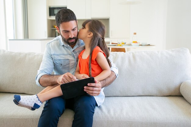 Adorable girl kissing her dad while he using tablet.