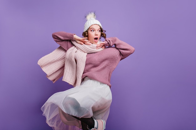 Adorable girl in trendy white hat fooling around during photoshoot. Indoor photo of blissful european woman in soft sweater and stylish long scarf.