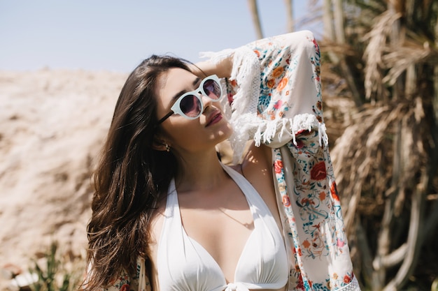 Adorable girl with dark hair wearing stylish bikini posing with eyes closed in sunny day. Attractive tanned brunette woman in sunglasses and swimsuit resting outside in vacation